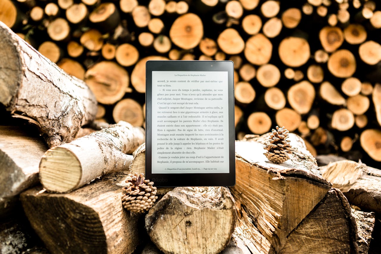 E-reader placed on a pile of wooden logs surrounded by pine cones outdoors.